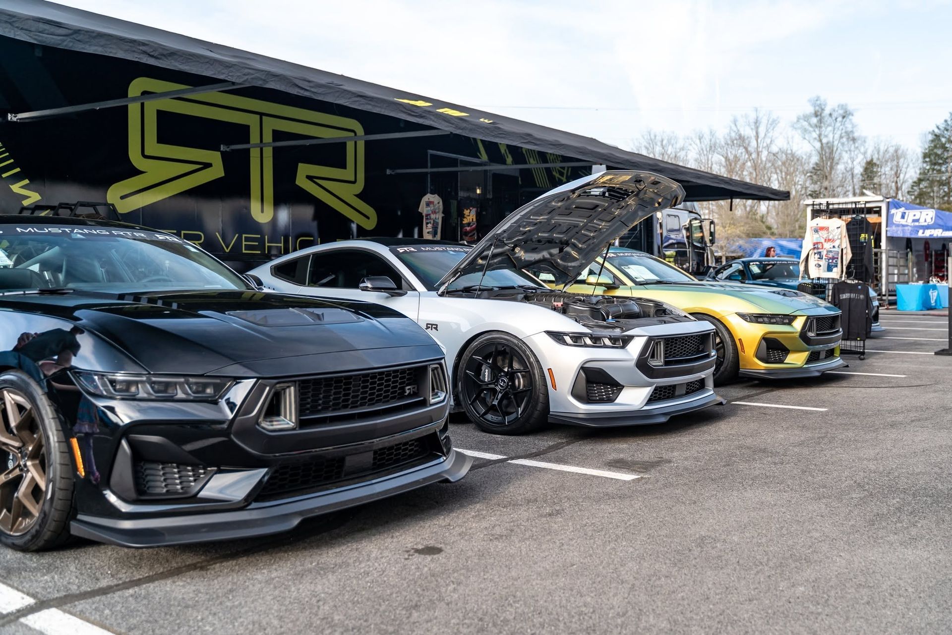 Row of customized sports cars at an outdoor car show, including black, white, and yellow vehicles with open hoods.