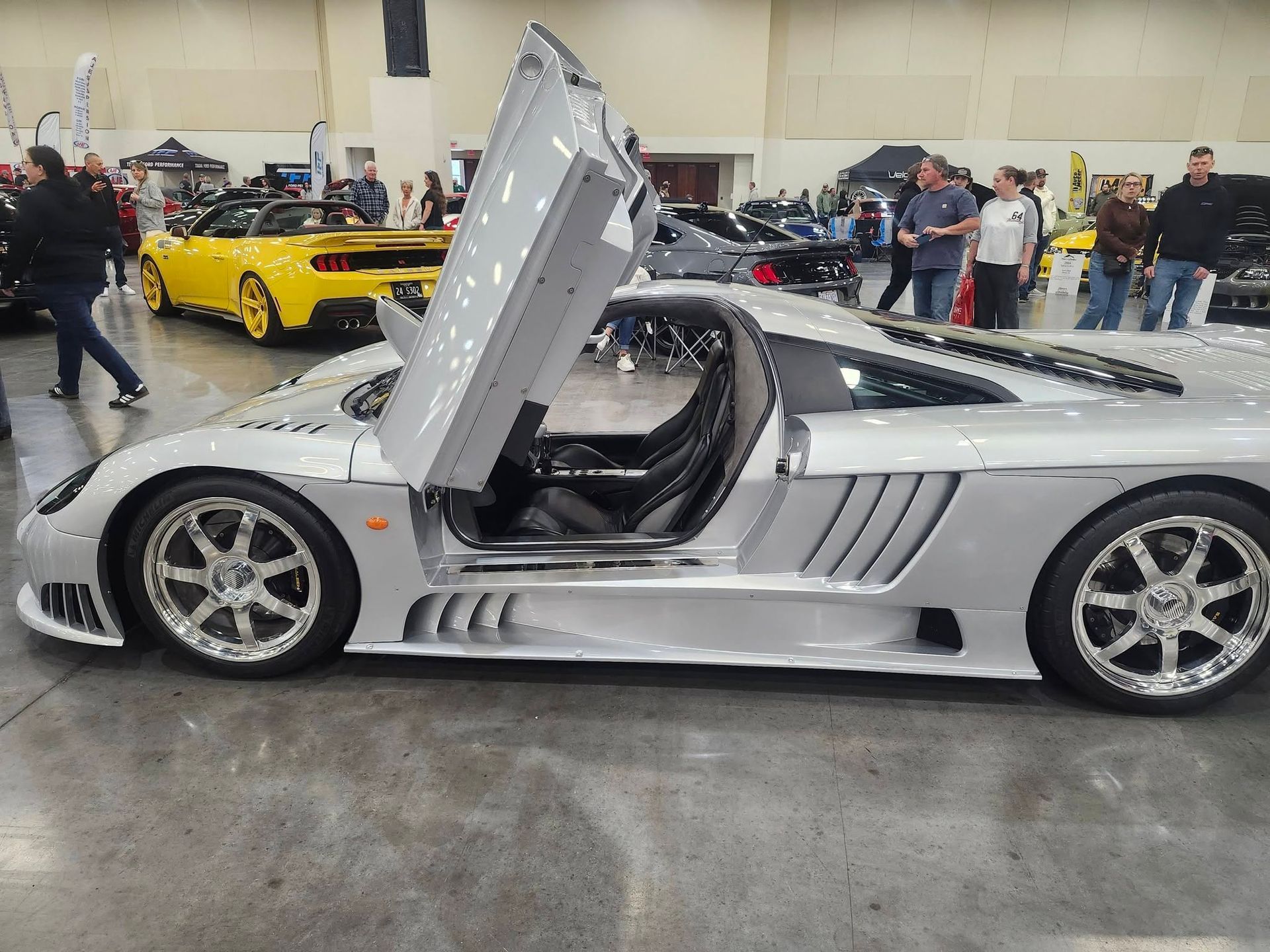 Silver sports car with scissor door open at an indoor auto show, with a yellow car in the background