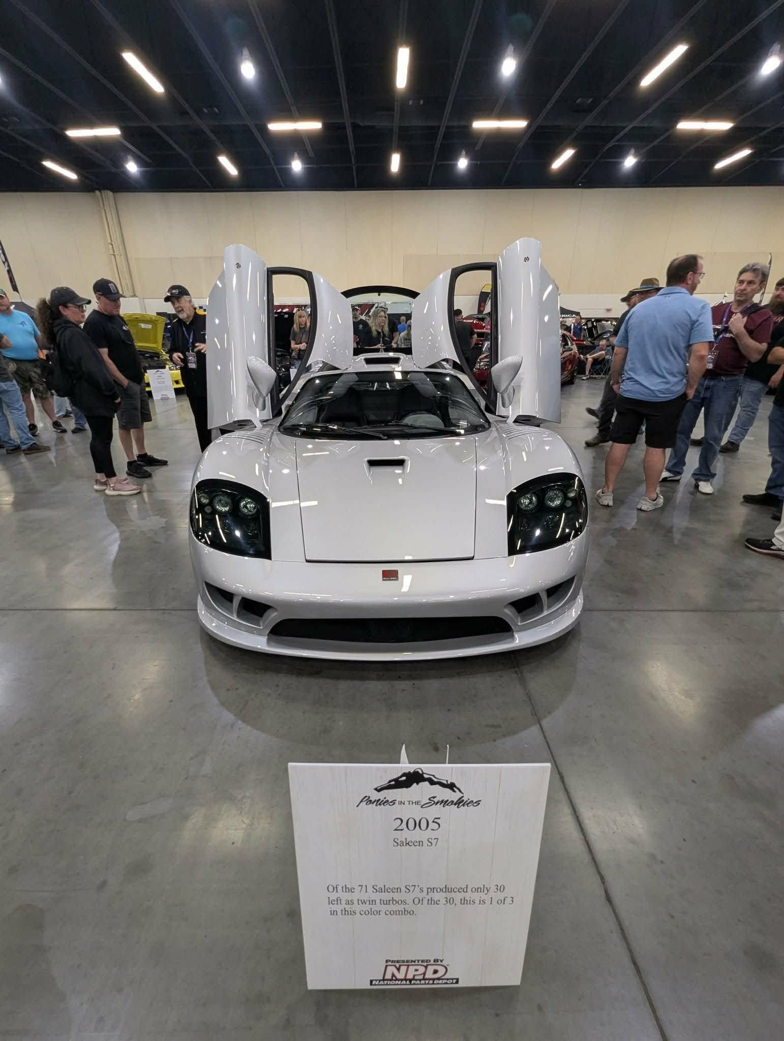 White sports car with butterfly doors open at an indoor auto show, surrounded by onlookers.