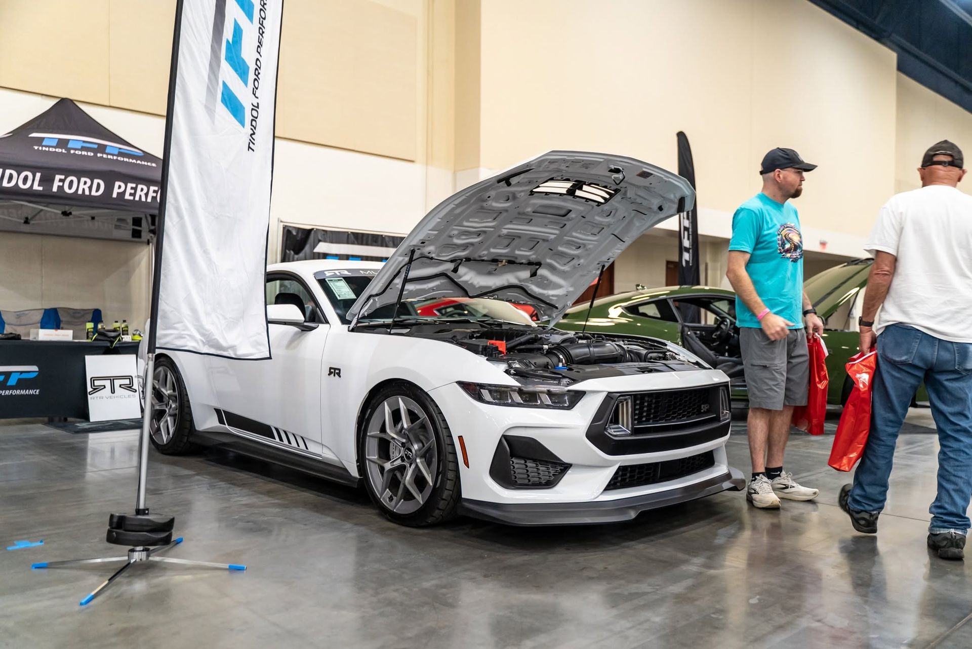 White sports car with hood open at an indoor auto show, with two men standing nearby.
