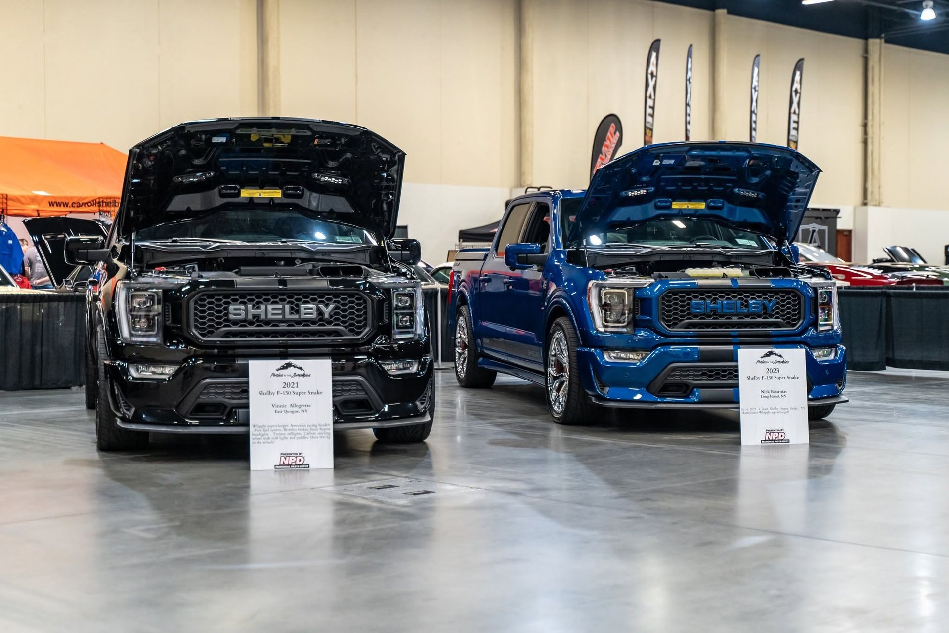 Two customized pickup trucks on display in a showroom, one black and one blue, with hoods open.