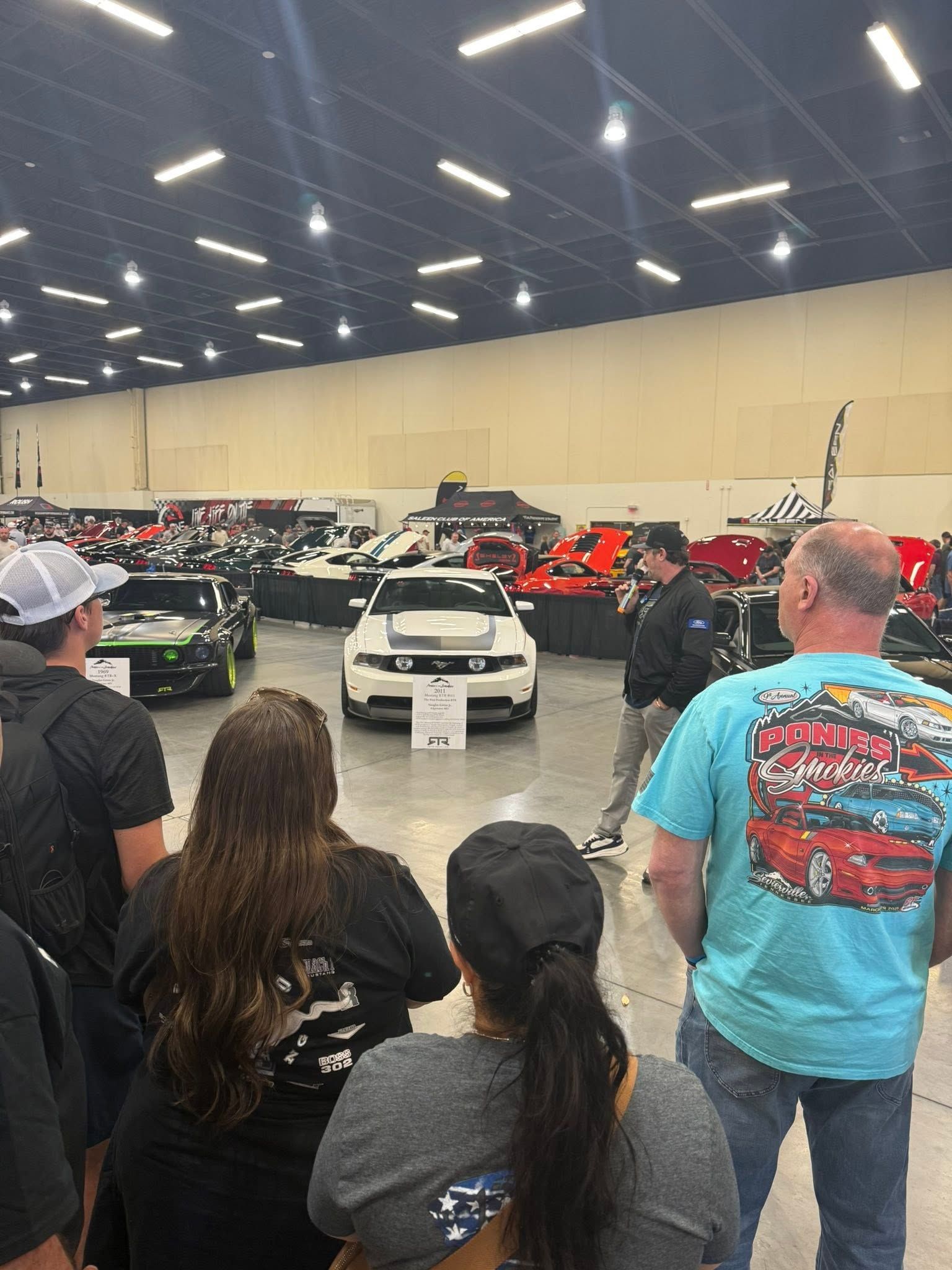 Crowd viewing rows of customized cars in a large indoor showroom, with a white car centered ahead.