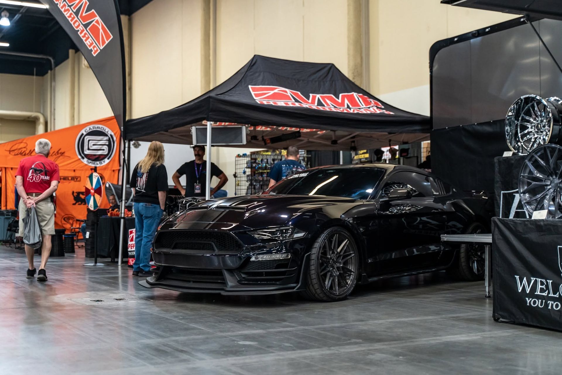 Black sports car on display at an indoor car show under a ZENKI tent, with people nearby.