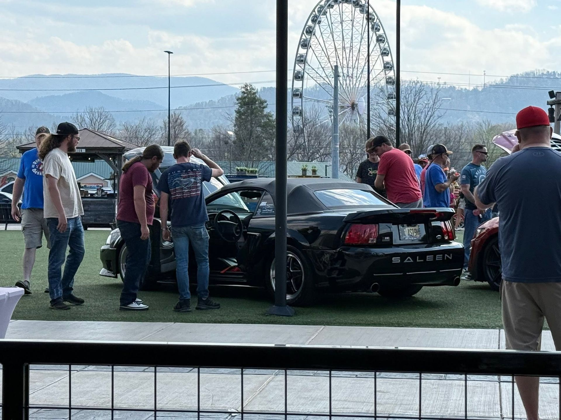 People gathered around a black sports car at an outdoor fair, with a ferris wheel in the background.