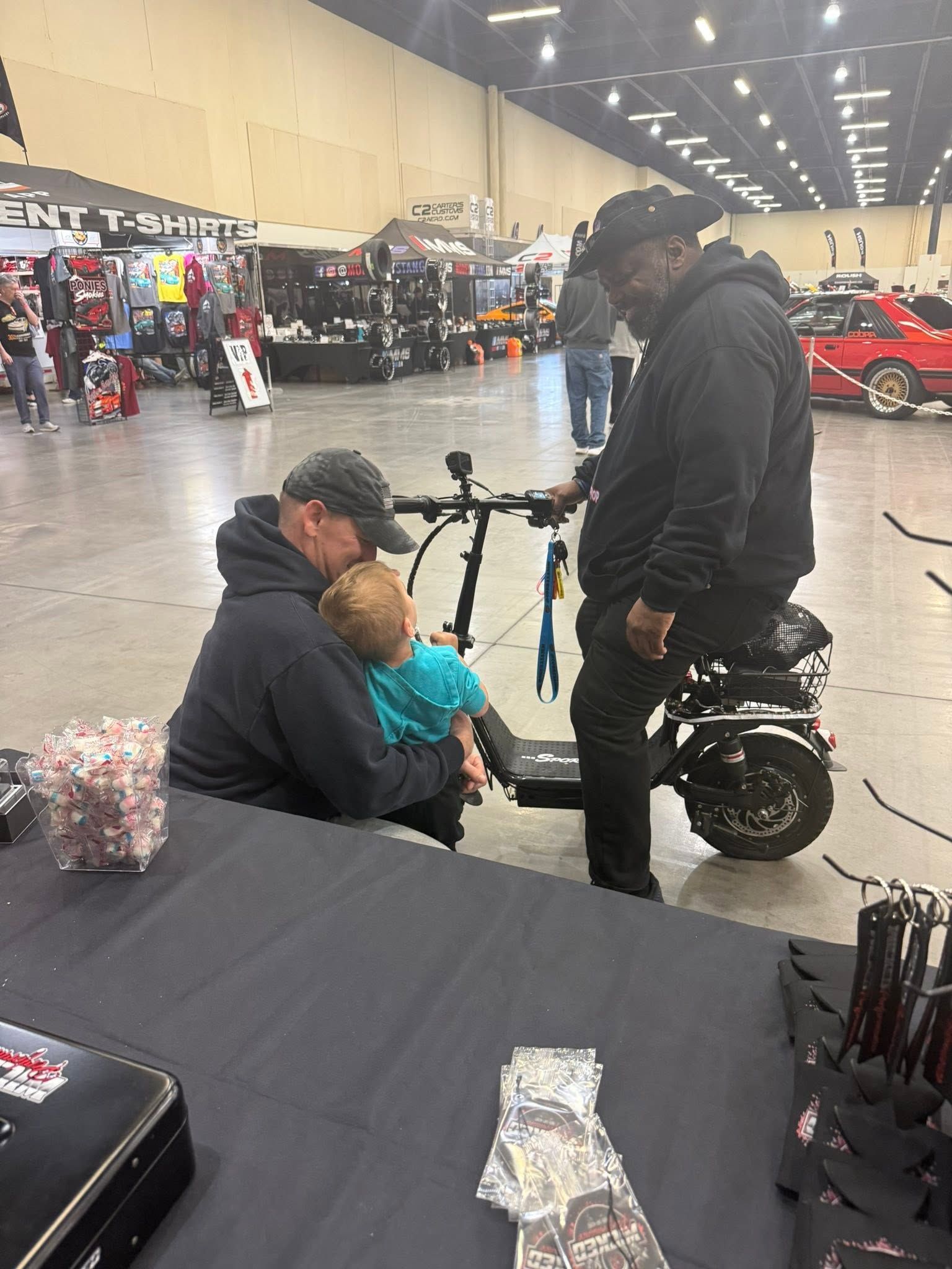 Man fitting a child onto a black ride-on scooter at a busy indoor expo hall with booths and vehicles nearby