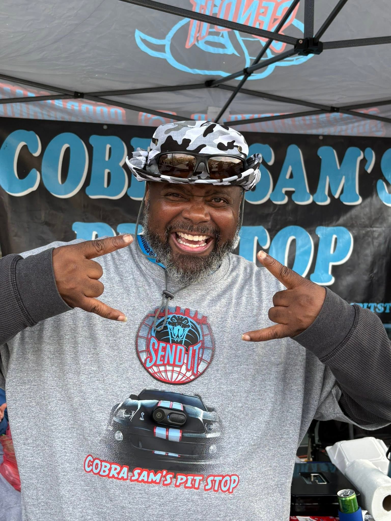Smiling man at a food booth, pointing at his shirt and wearing a cap under a canopy.