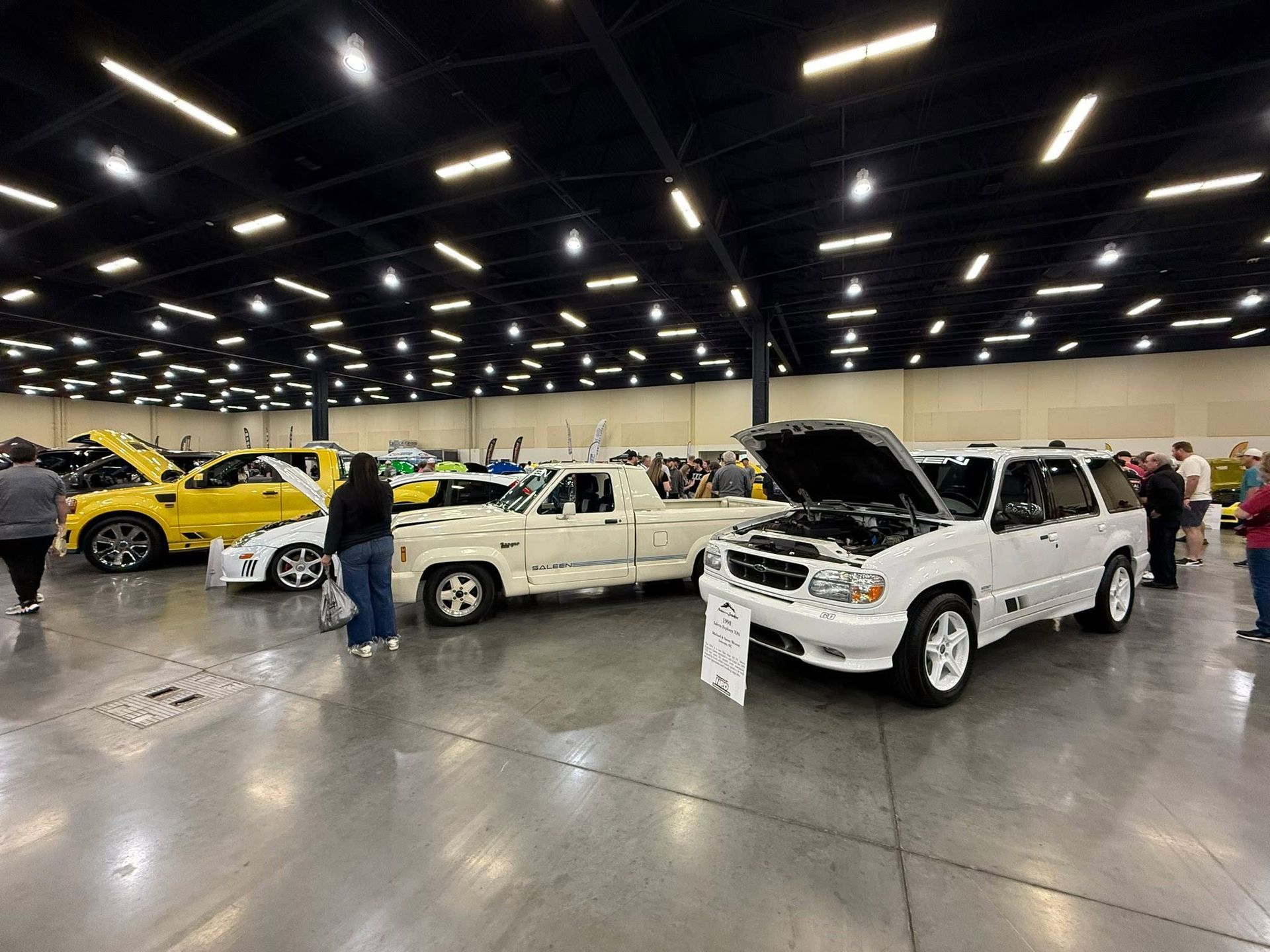 Classic cars displayed in an indoor showroom, including a white pickup with its hood open and a yellow sports car.