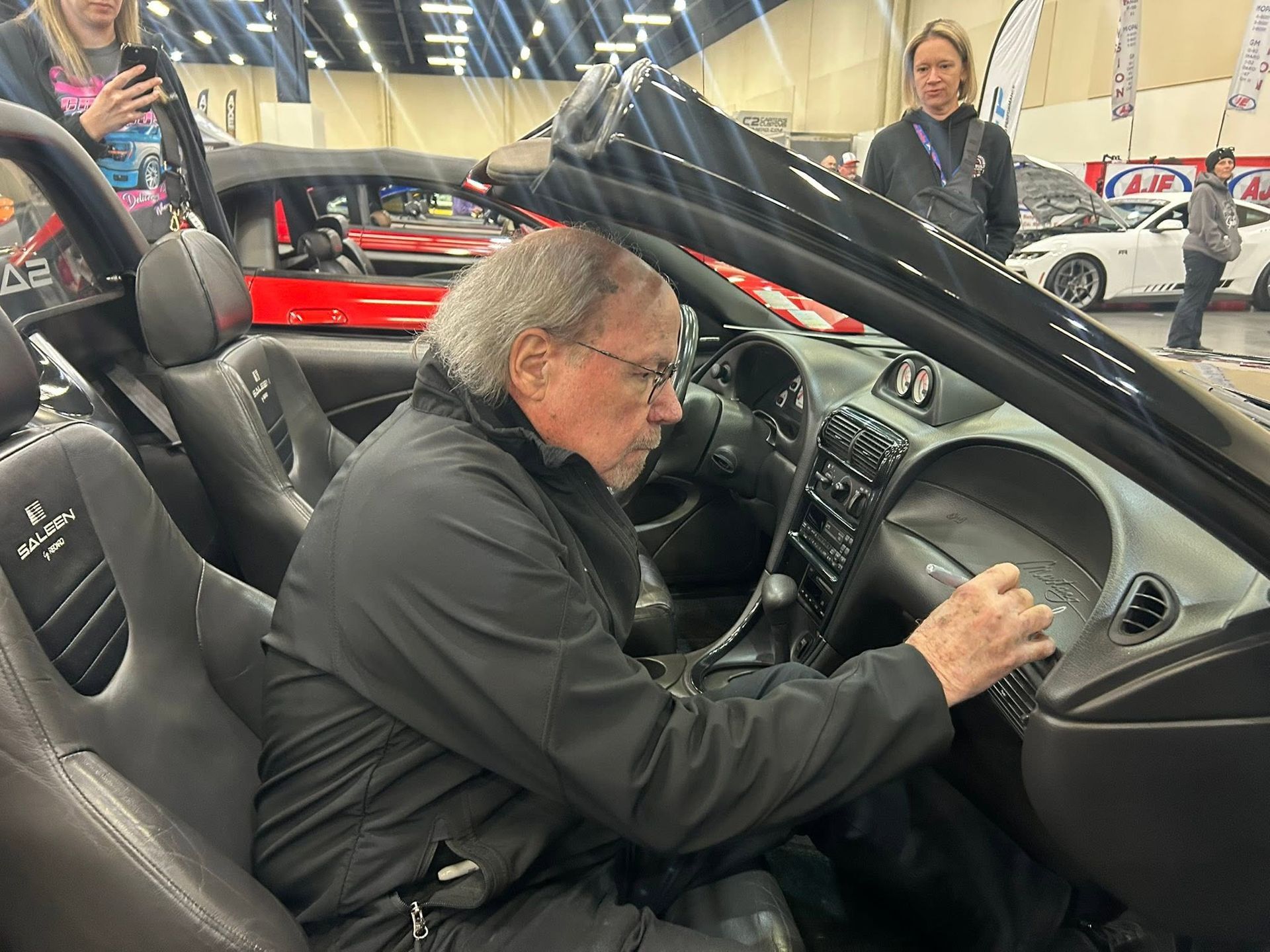 Man sitting in a black sports car, adjusting the dashboard at an auto show.