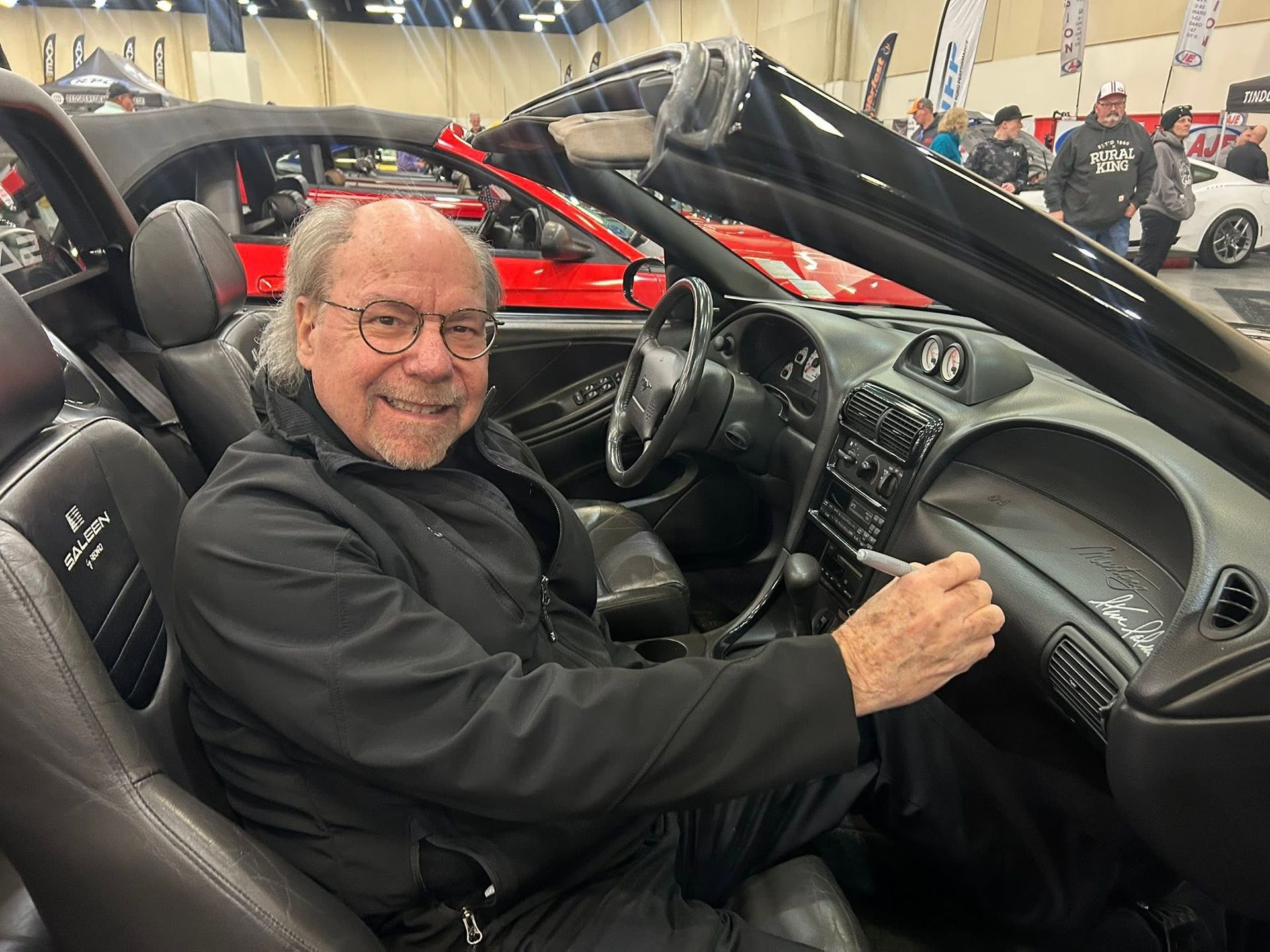 Smiling man sitting in a black sports car at an indoor auto show