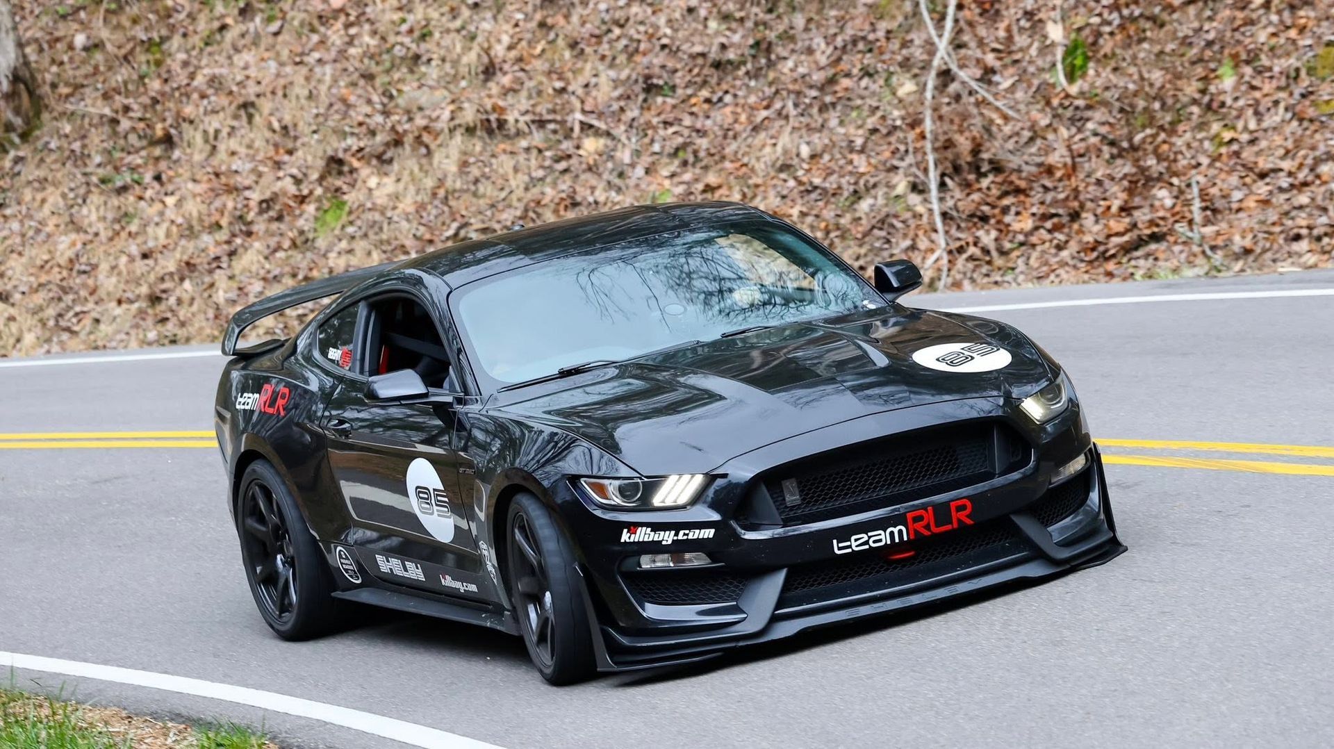 Black sports car with decals driving on a winding road beside a wooded hillside