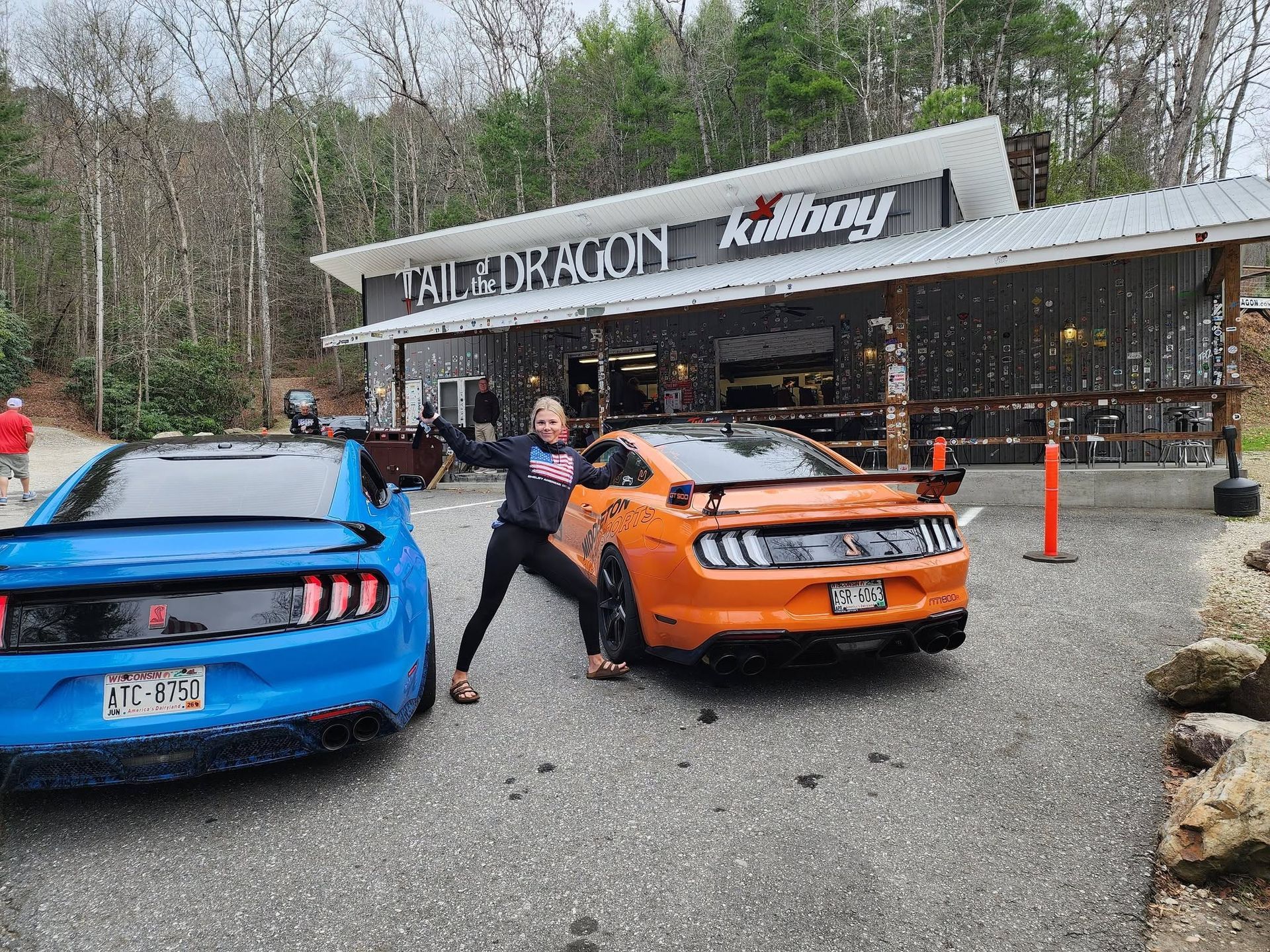 Person posing between blue and orange sports cars outside a café.