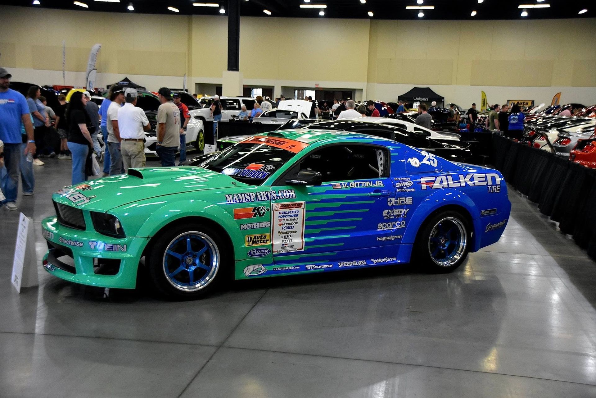 Green-and-blue race car displayed indoors at an auto show, surrounded by spectators and other cars.