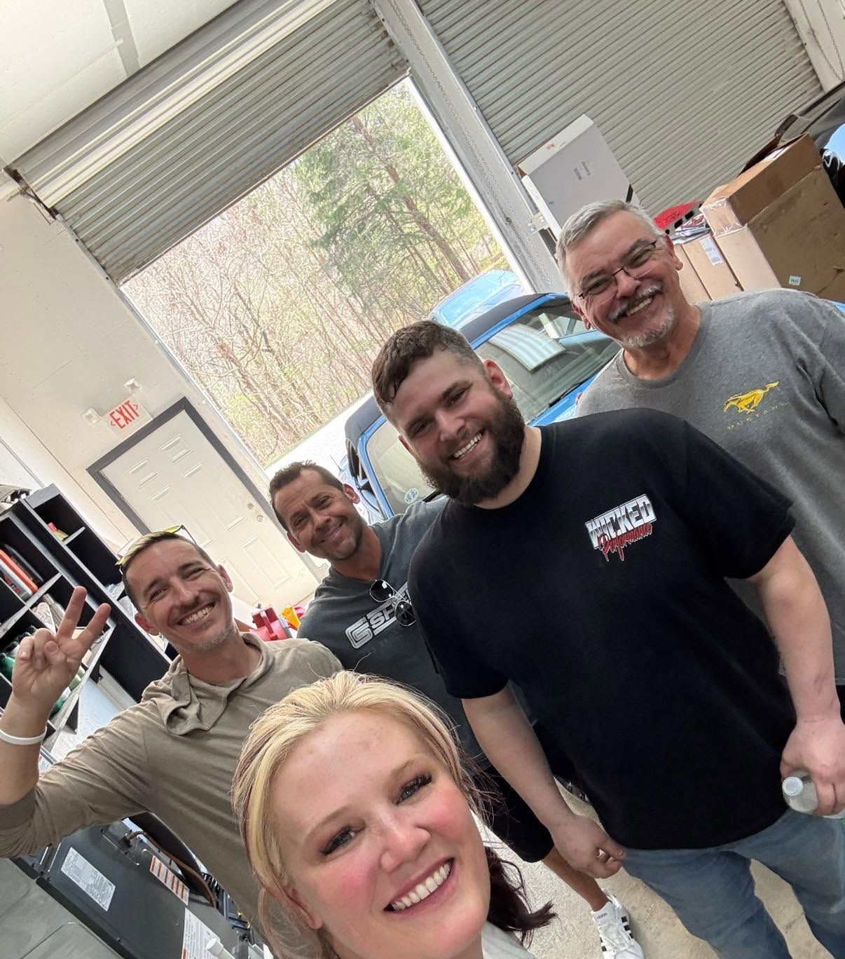 Group selfie of five smiling coworkers in a workshop, one waving at the camera.