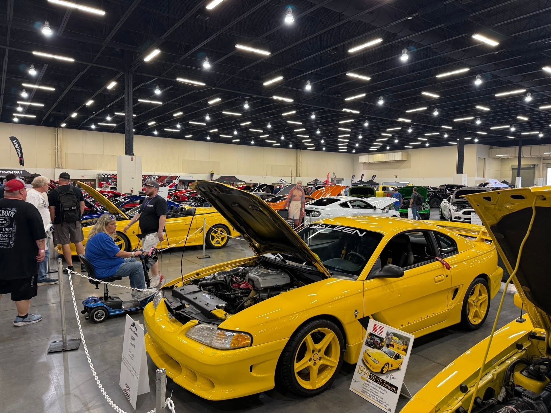 Yellow sports cars displayed indoors at a car show, with one car’s hood open in the foreground.