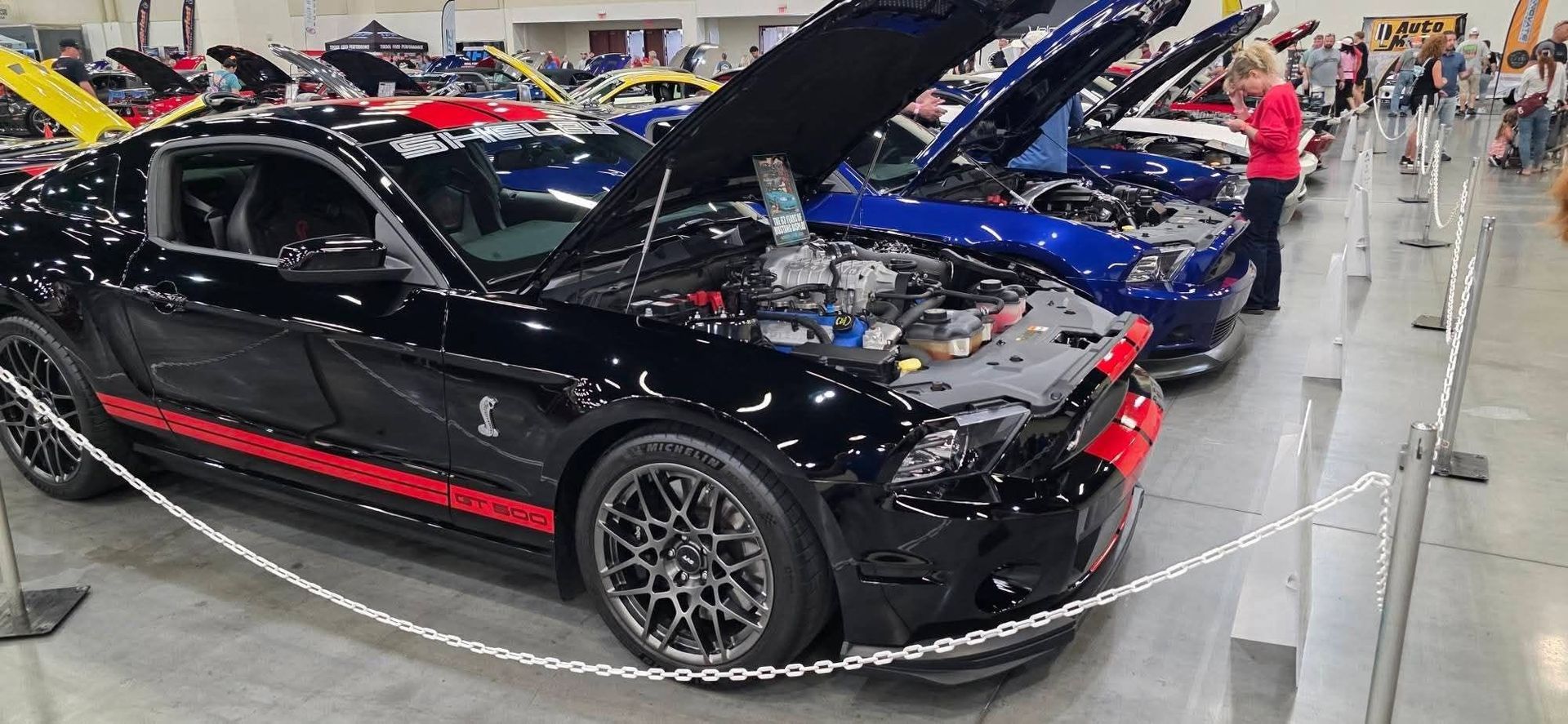 Black sports car with hood open on display at an indoor car show, surrounded by other custom cars