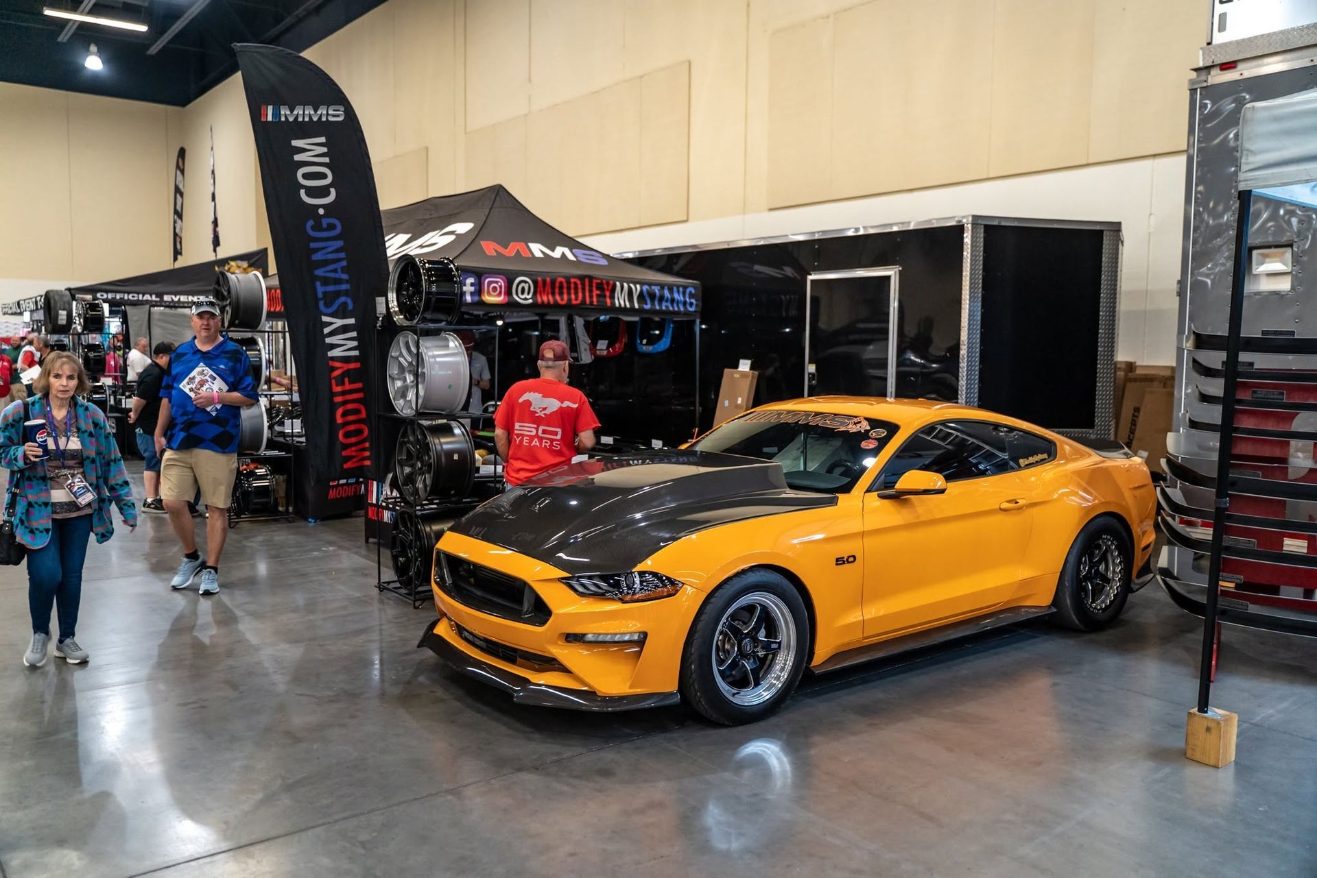 Orange sports car on display at an auto show, with booths and attendees in the background