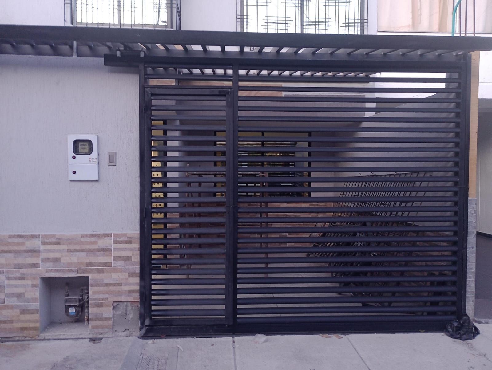 Puerta metálica negra y toldo que cubre la entrada. Edificio con paredes claras, ladrillo beige y cajas de servicios públicos.
