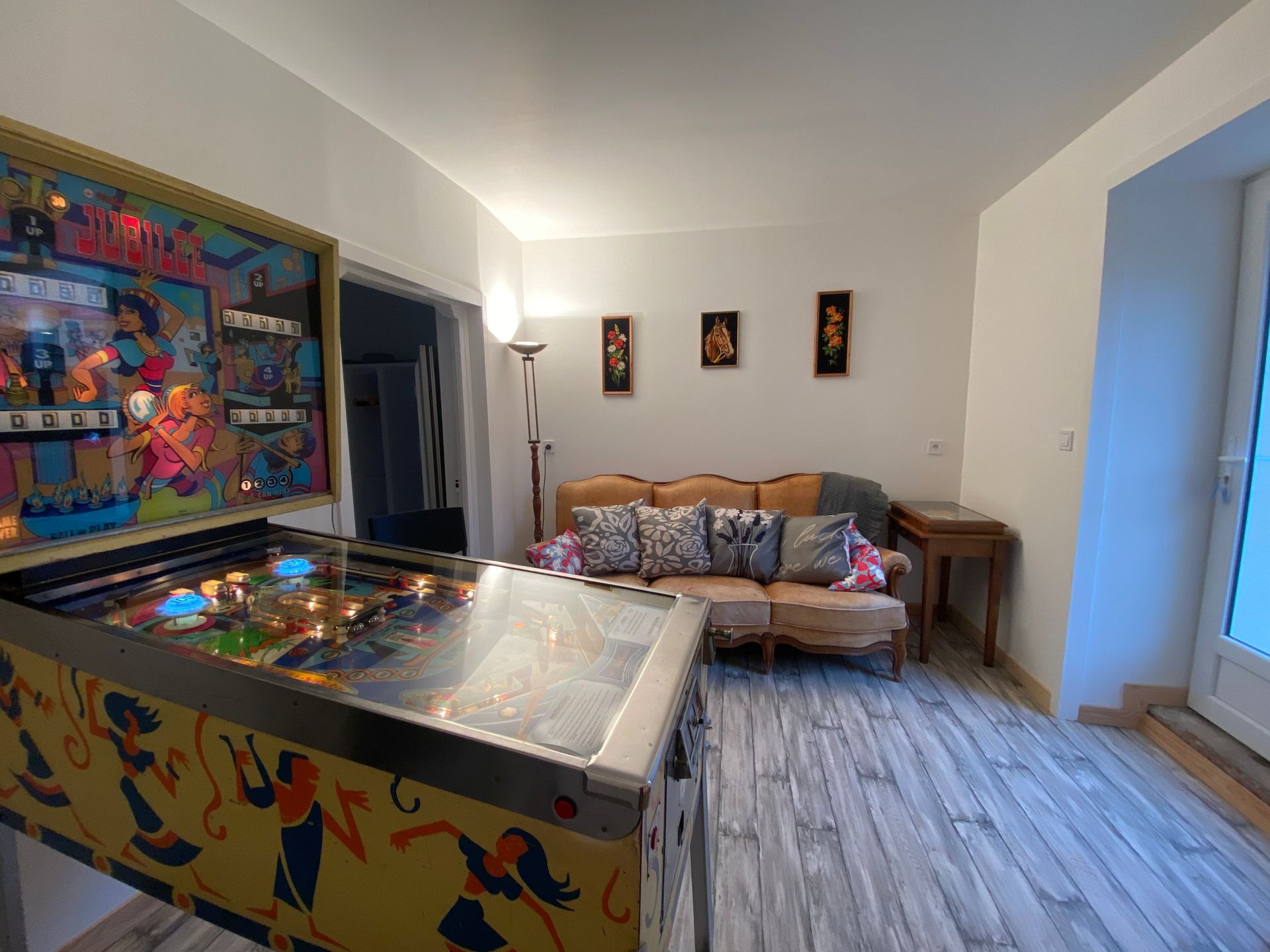Un salon , flipper, un canapé jaune usé avec des coussins, trois cadres et une porte baignée de lumière. 