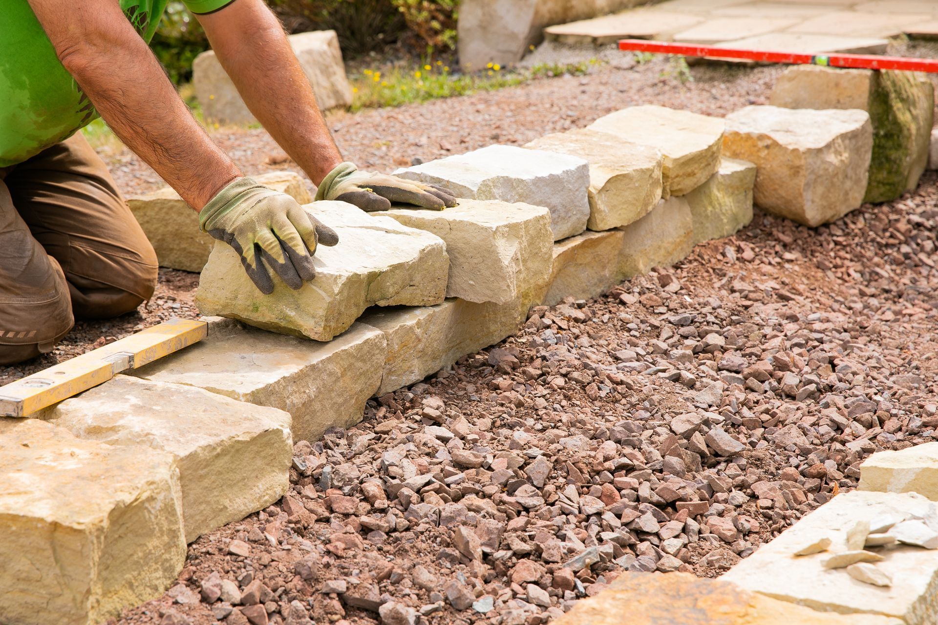 Person building a stone wall, wearing gloves, in a garden setting, placing a large stone.