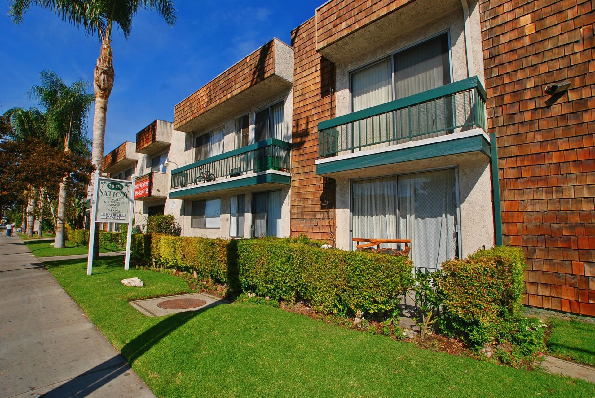Apartment building with brown shingled roof, balconies, and green shrubs along a sidewalk.