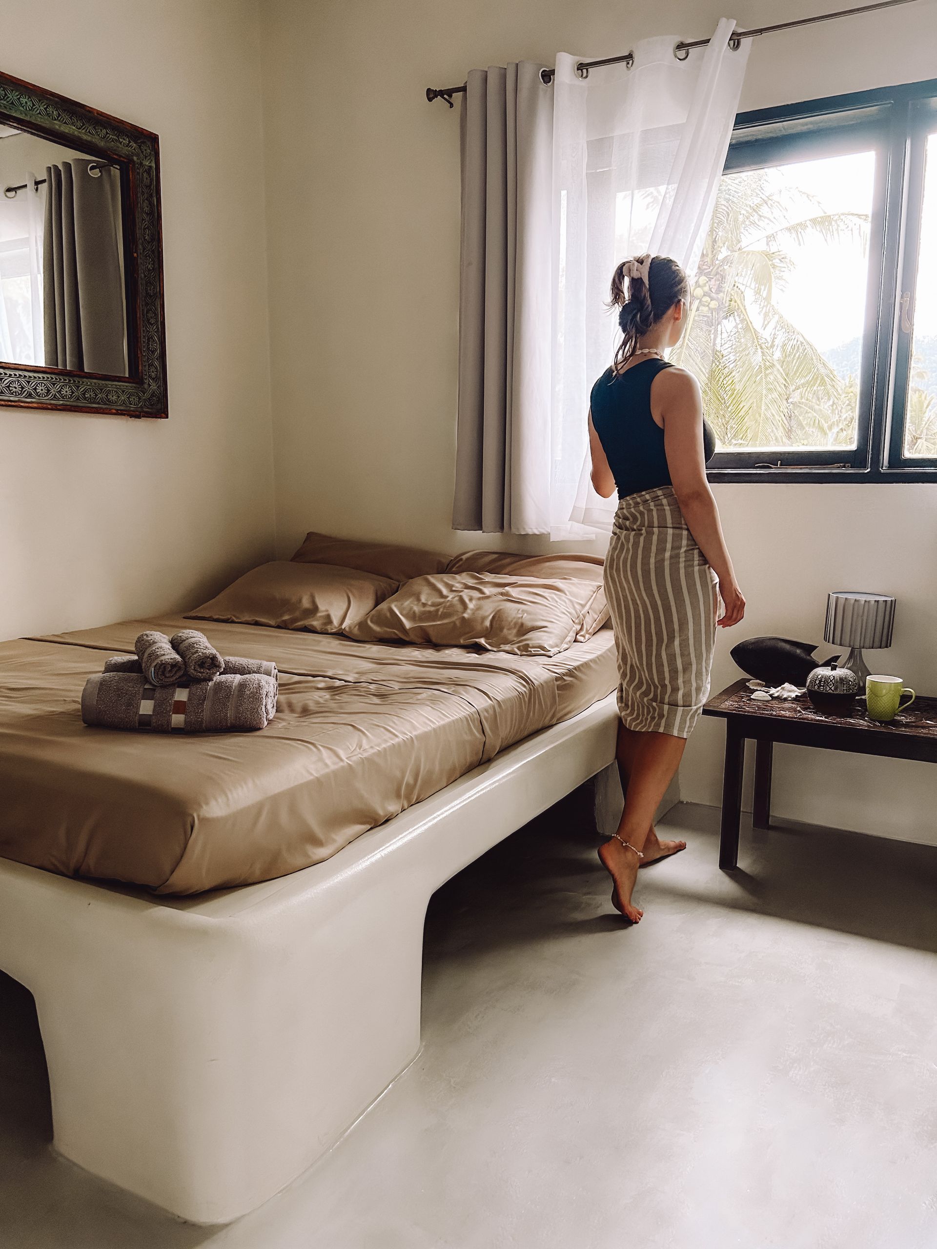 A woman is standing in a bedroom next to a bed looking out of a window.