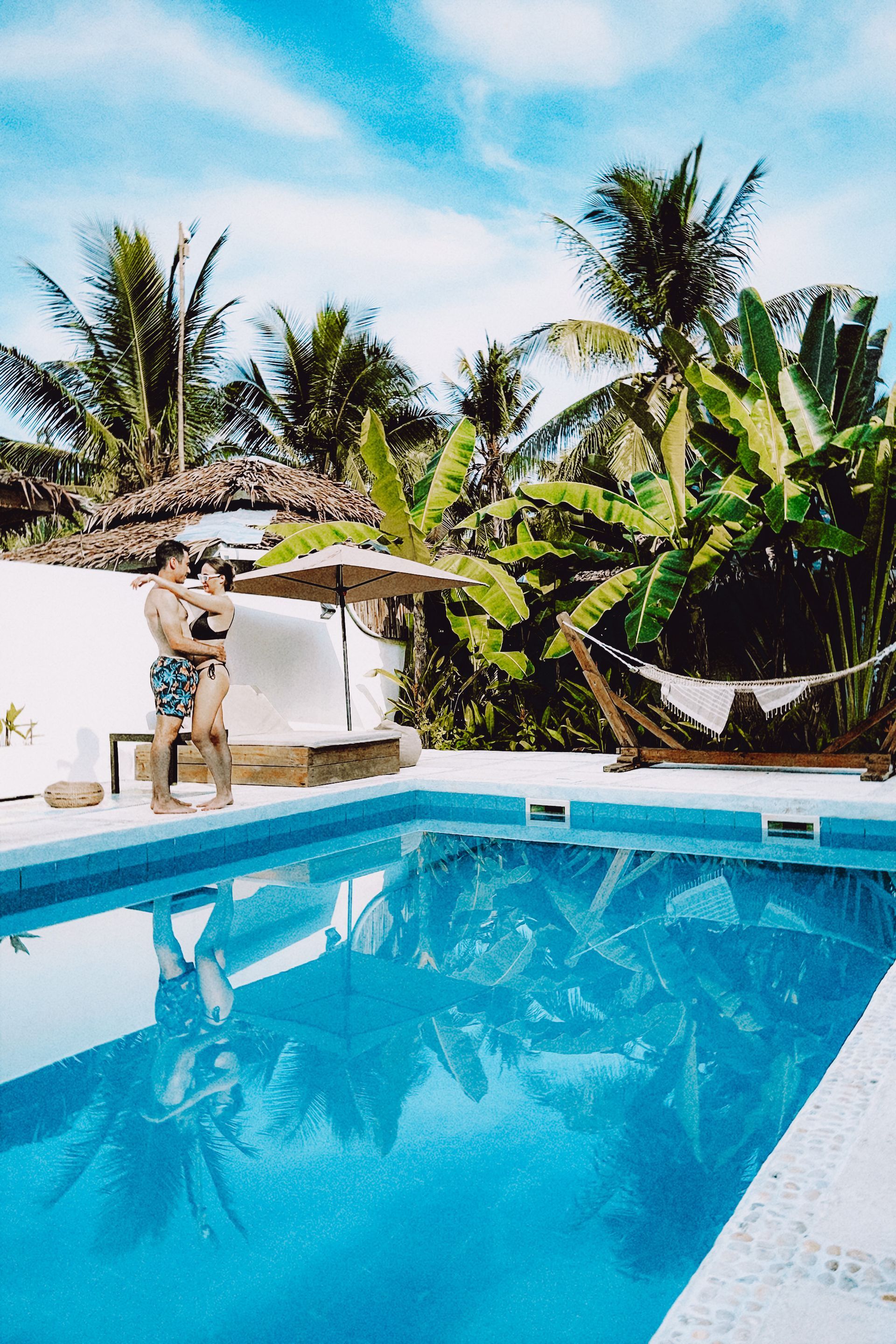 A man and woman are standing next to a swimming pool surrounded by palm trees.