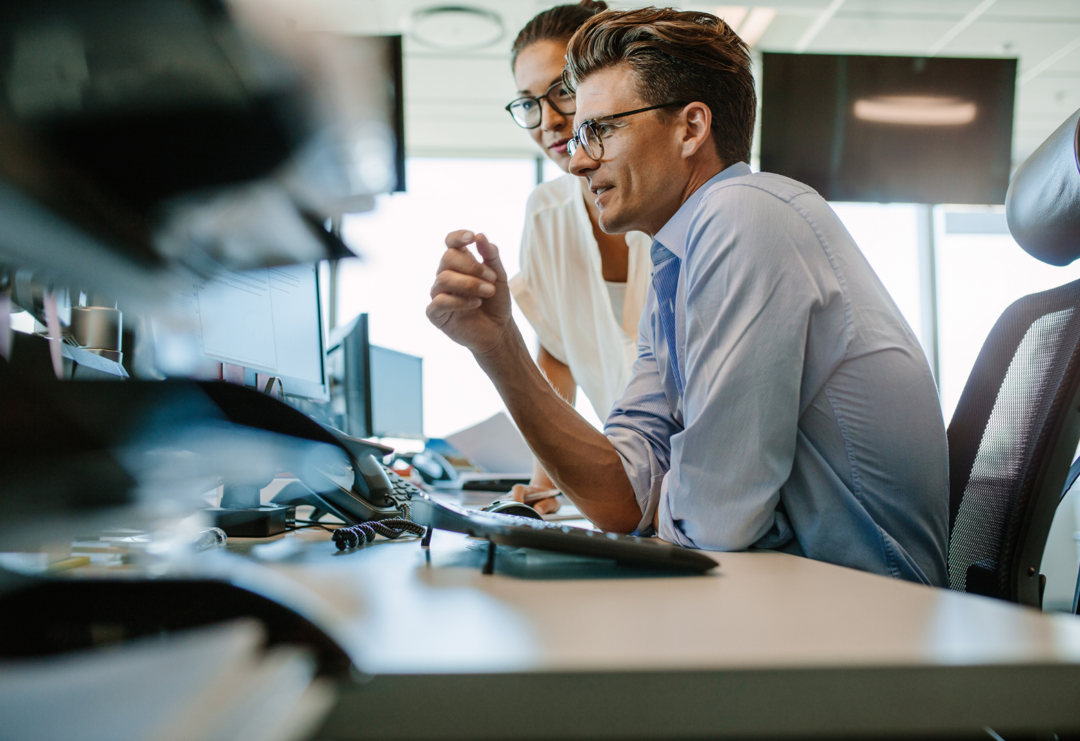 Two coworkers at a desk reviewing work on a computer in a bright office