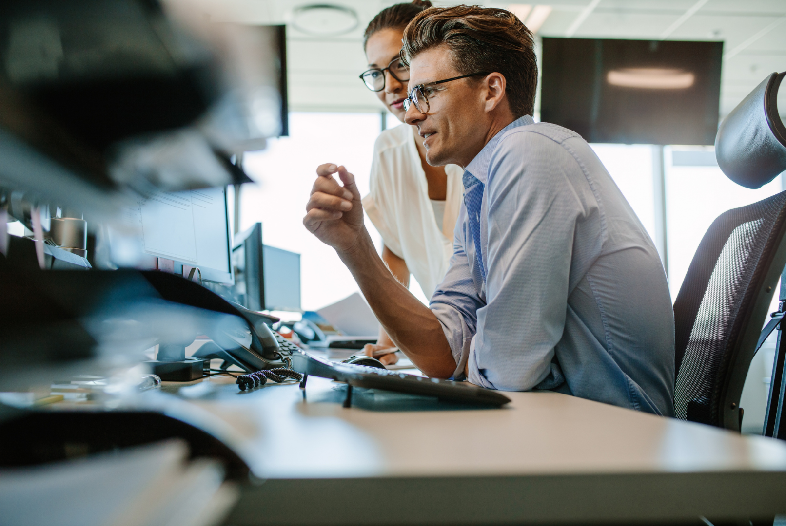 Two coworkers reviewing data at a desk with multiple monitors in an office