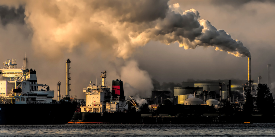 A large ship is floating in the water next to a factory with smoke coming out of the chimneys.