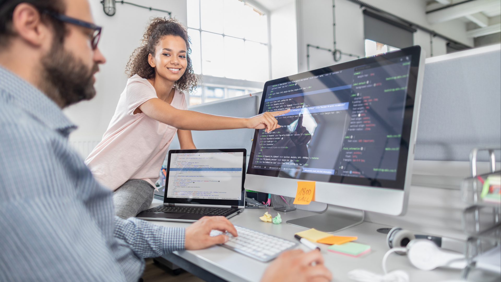 Two coworkers in a bright office pointing at code on a large monitor and a laptop