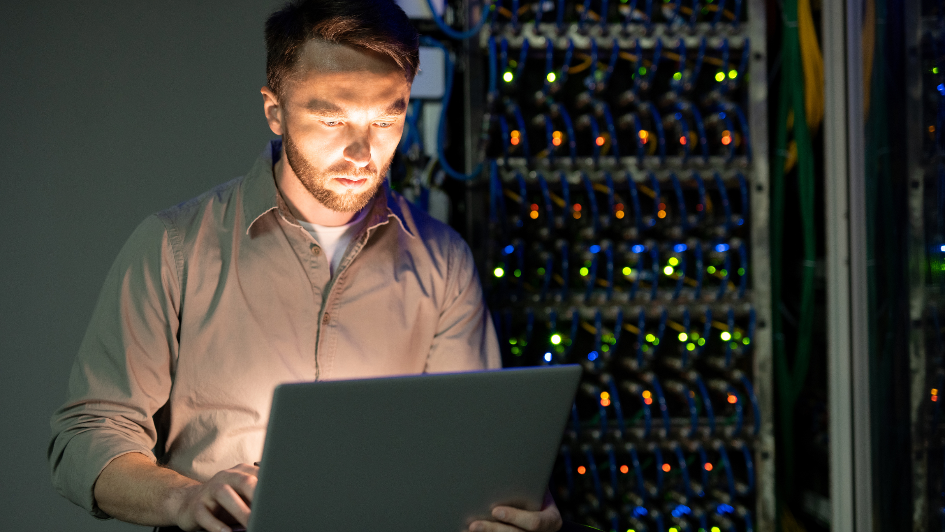 Technician working on a laptop in a server room, illuminated by screen light and surrounded by server racks.