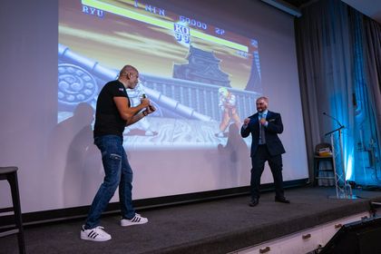 Two men are standing on a stage in front of a large screen playing a video game.