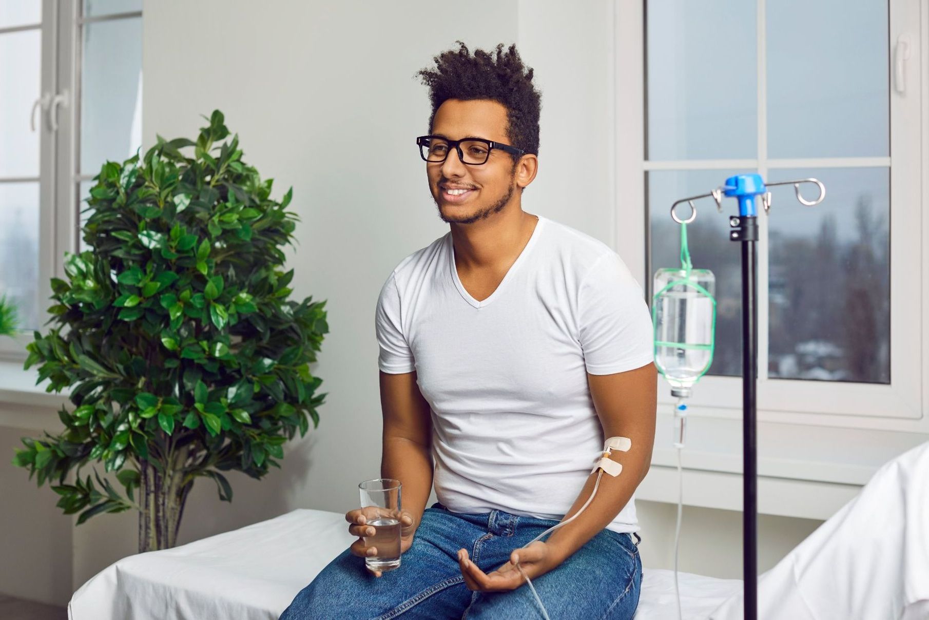 A man is sitting on a bed with an iv in his arm holding a glass of water.