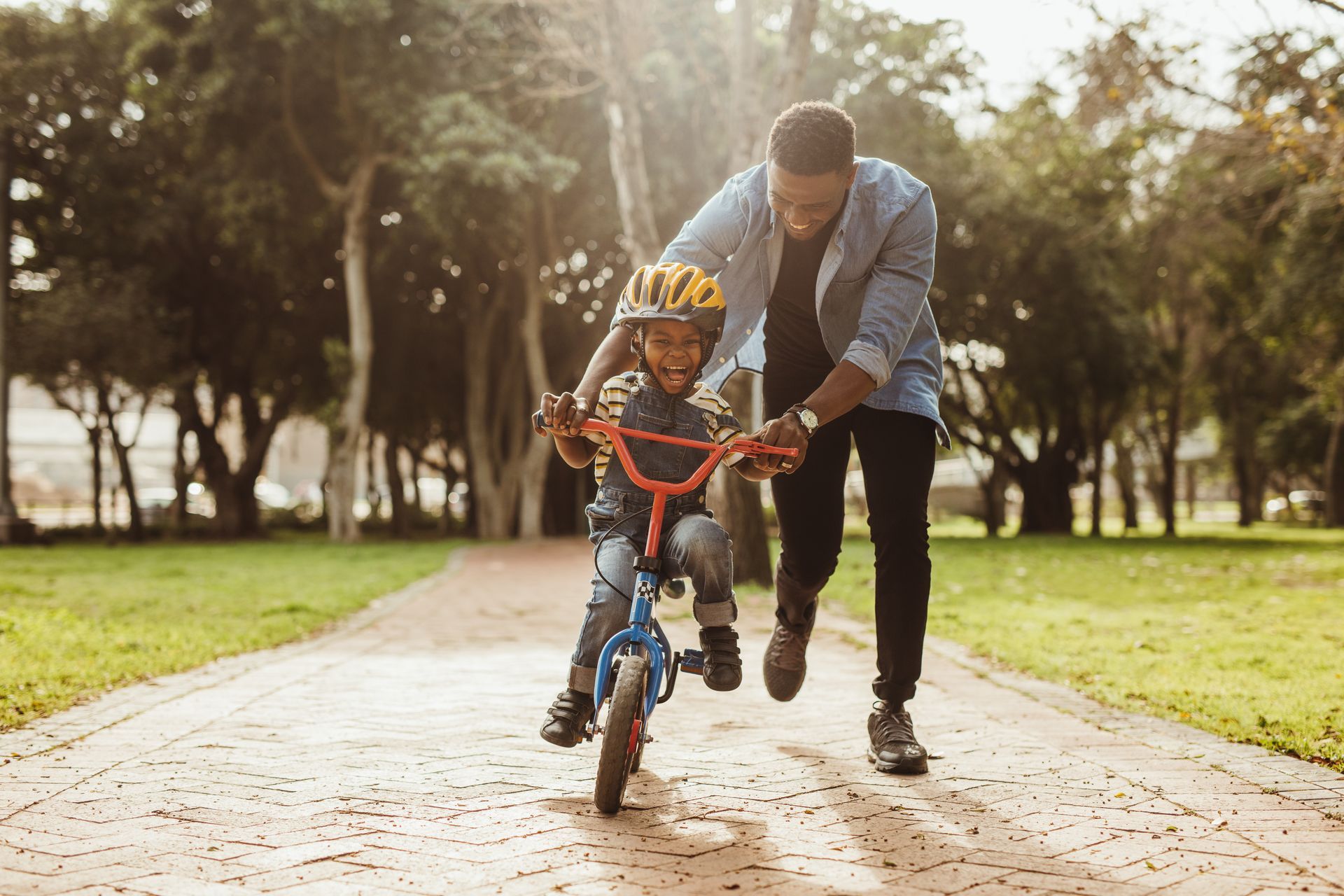 A father helps his smiling child ride a blue bike in a park.