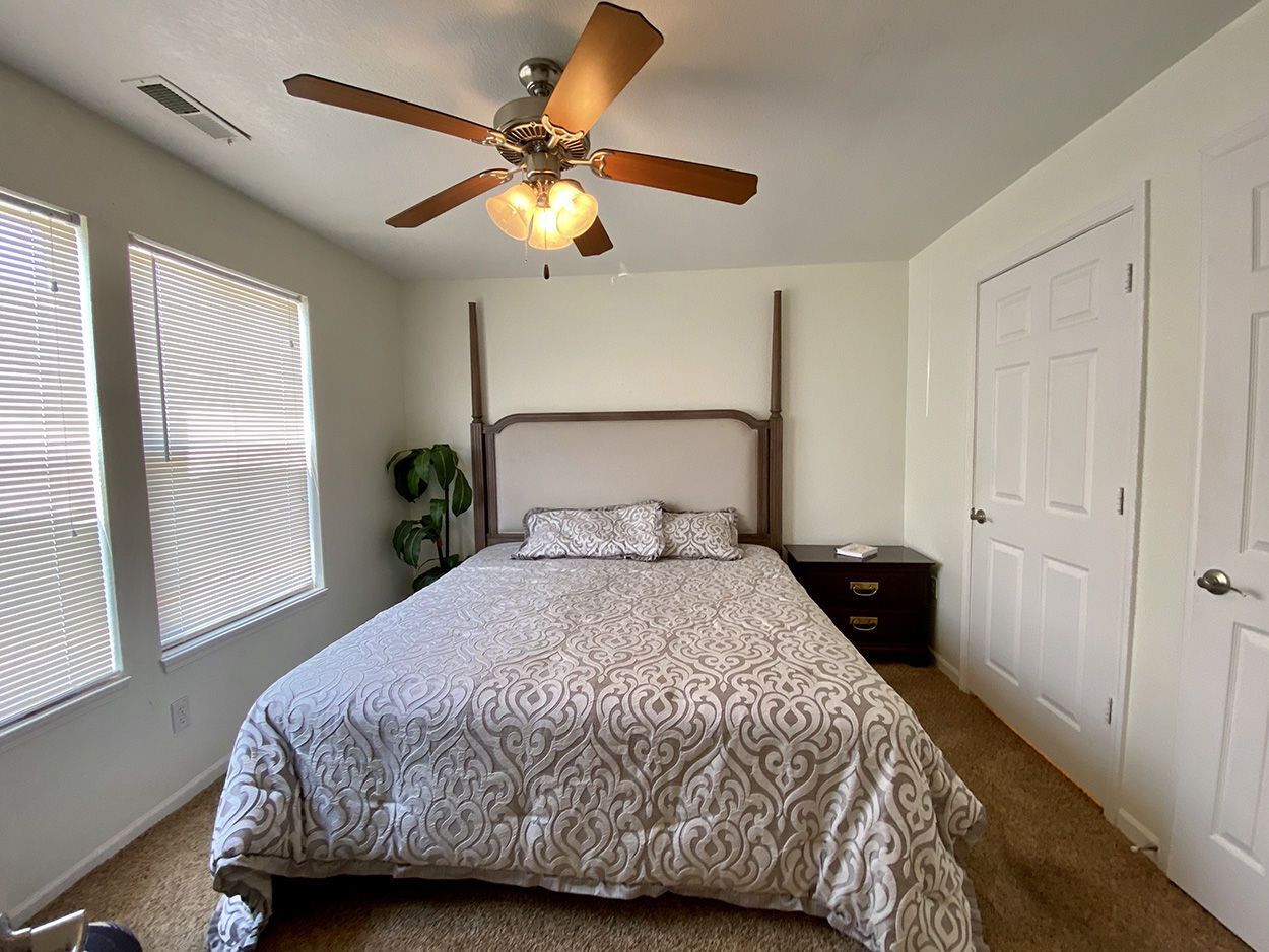 Bedroom with bed, nightstand, windows with blinds, and ceiling fan.