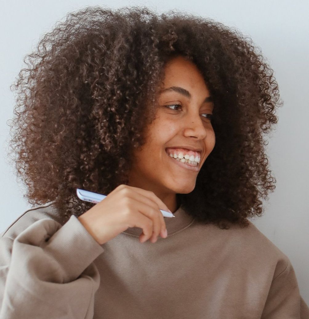A woman with curly hair is brushing her teeth and smiling.