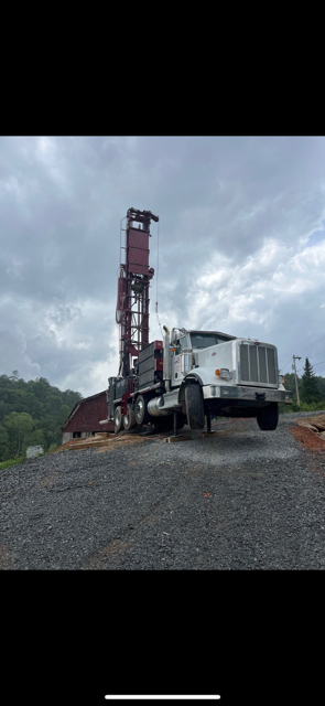 White drilling truck beside a tall red drilling rig on a gravel site under cloudy skies