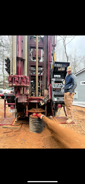 Drilling rig boring into red soil as a worker stands nearby at a muddy site