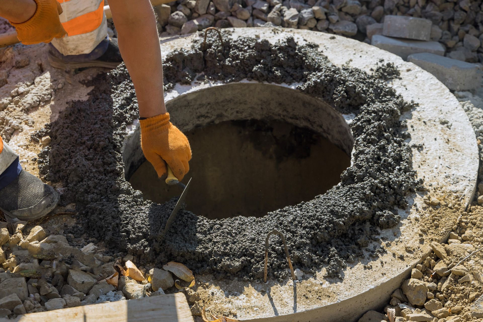 A worker in orange gloves trowels wet concrete around a circular well casing ring at a job site