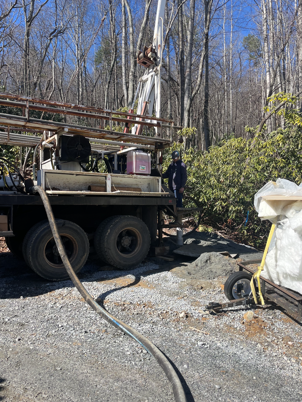 Utility truck with hose at a wooded construction site beside a gravel driveway