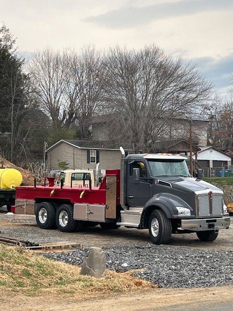 Gray dump truck parked on gravel in a rural yard, with red bed and leafless trees in the background