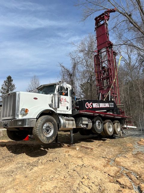 White flatbed truck carrying a tall red drilling rig on a dirt site under a blue sky