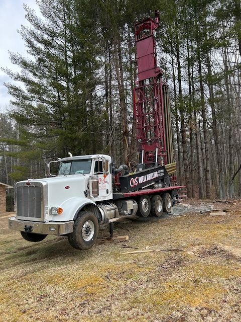 White drilling truck with red derrick parked in a wooded clearing on a gravel surface