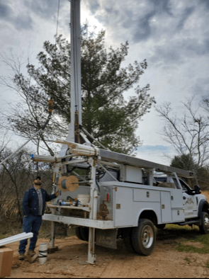 Utility truck with raised mast and worker standing beside it in a wooded outdoor area