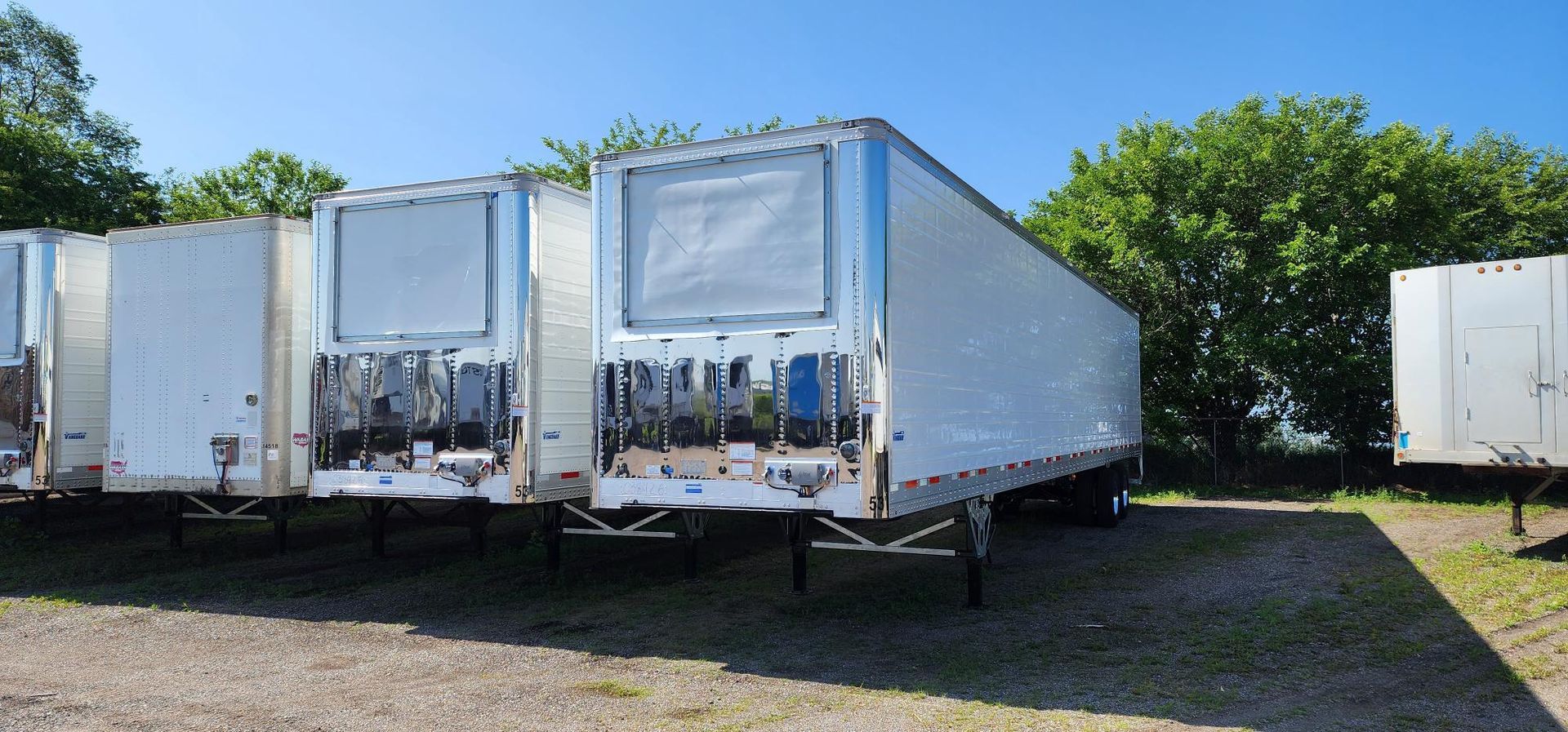 Several white semi-trailer trucks parked on gravel under a blue sky, surrounded by green trees.