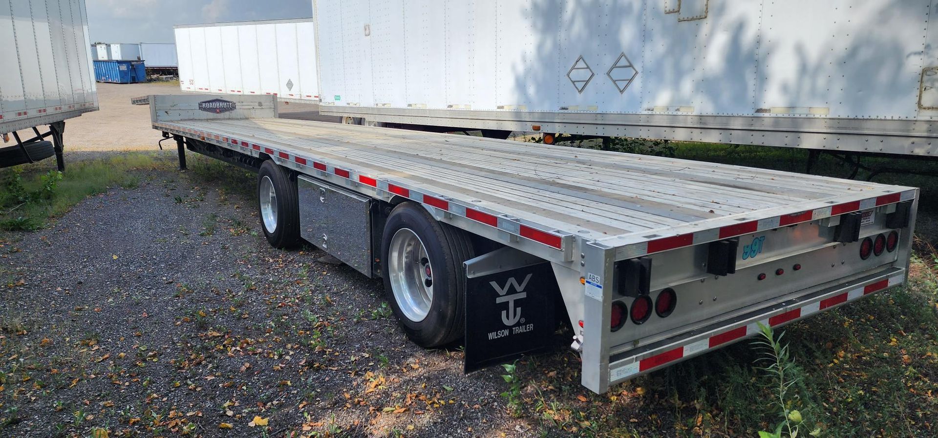 Flatbed trailer parked on gravel, with grass. White storage containers in the background.