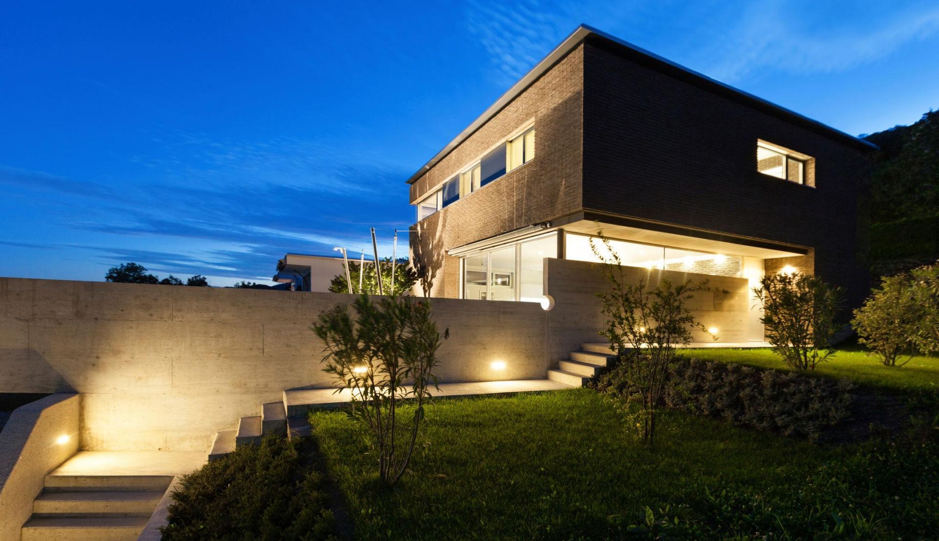 Modern two-story house at dusk with warm exterior lights and a lit stone wall along the front yard