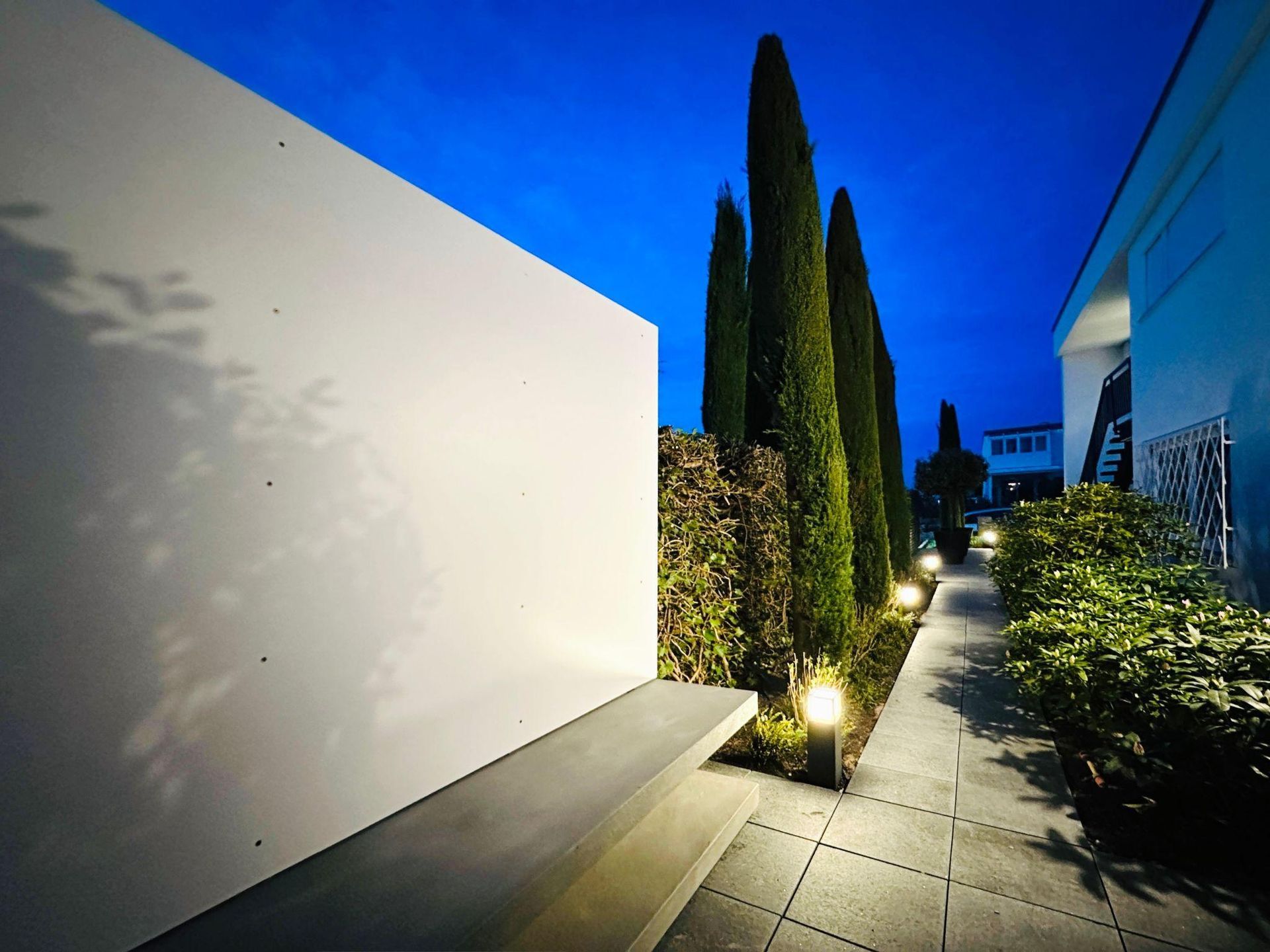 Narrow garden path beside white walls, lit by lamps and tall cypress trees at dusk.