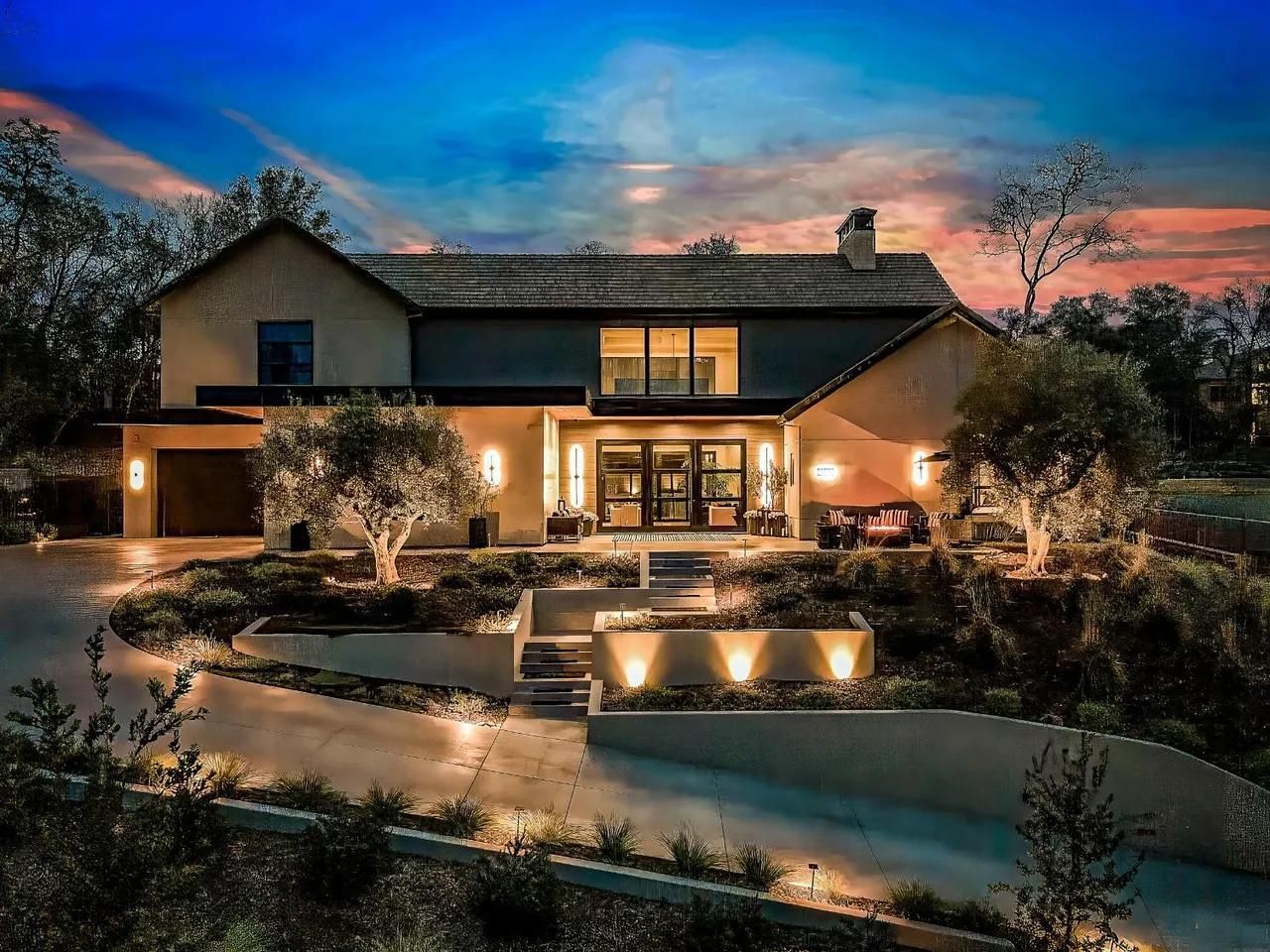 Modern two-story house at dusk with warm lights, landscaped garden, and a colorful sunset sky