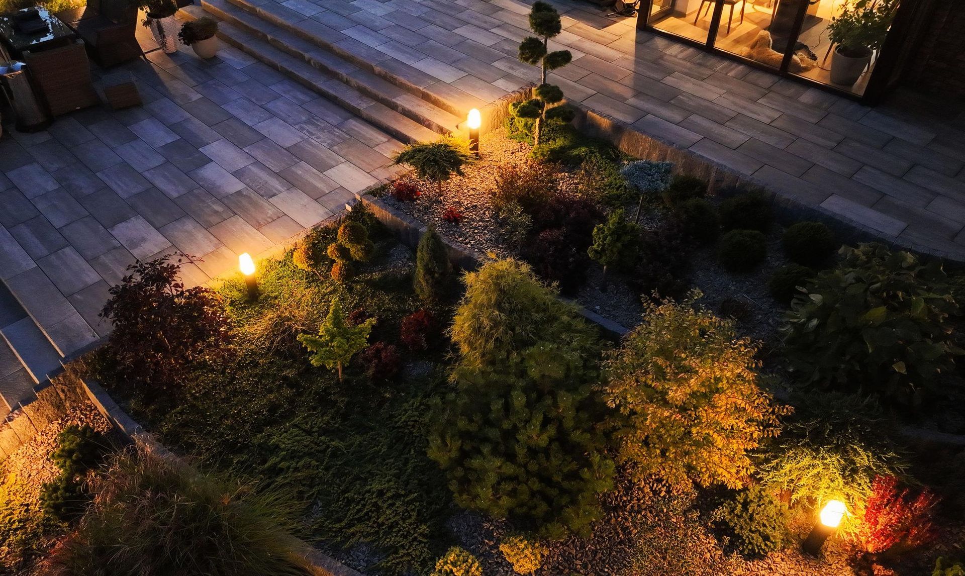Small landscaped garden at night, lit by warm path lights beside a paved walkway.